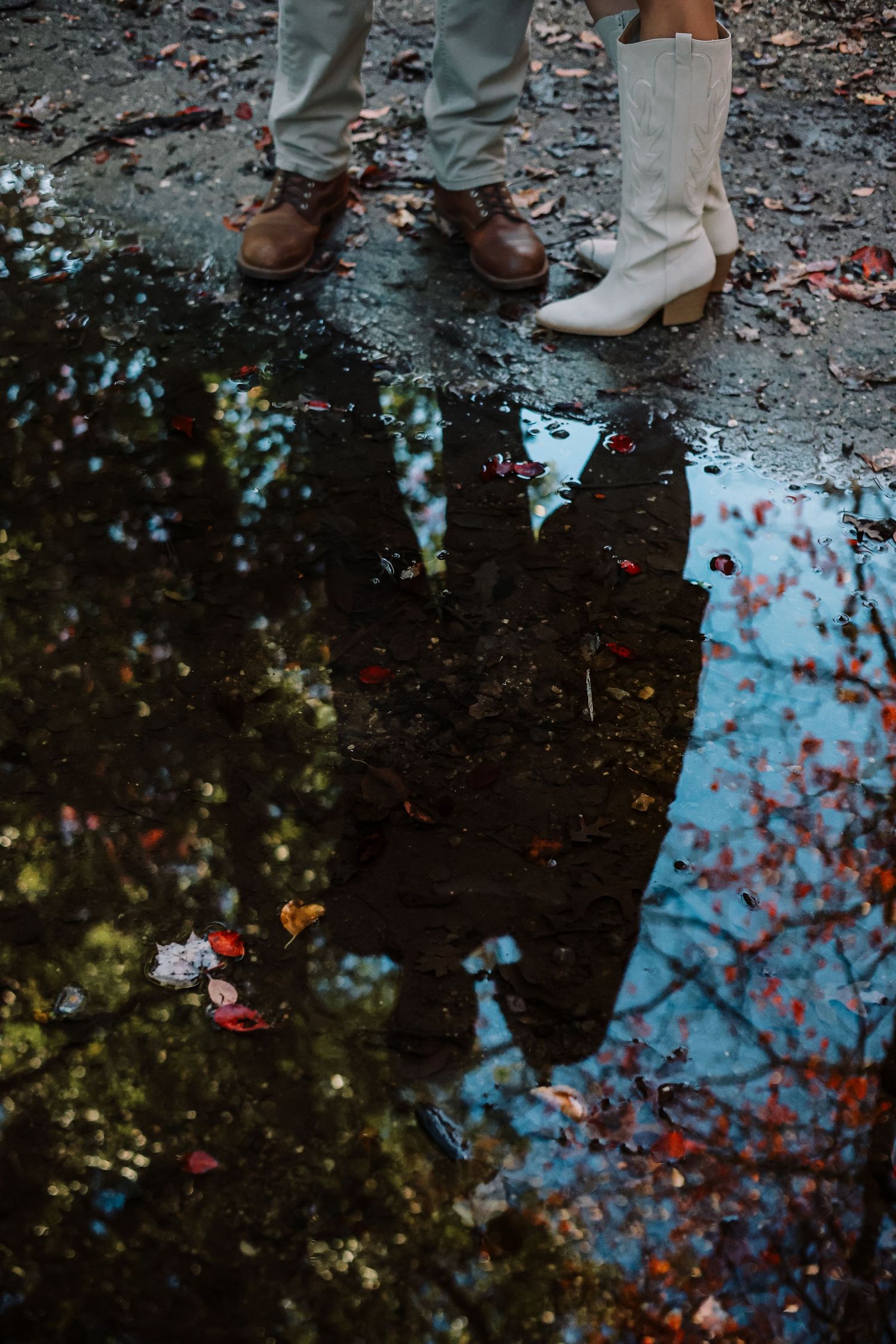 Reflection of a couple standing close together in a rain puddle surrounded by fallen autumn leaves, with boots visible above.