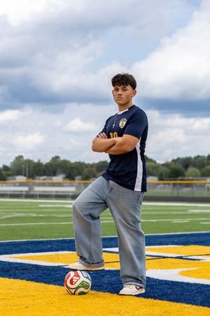 Teen soccer player in navy jersey poses with arms crossed, foot on ball, on a yellow and blue turf field.