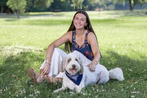 Young woman sitting on green grass in a park with a white fluffy dog wearing a blue bandana on a sunny day.