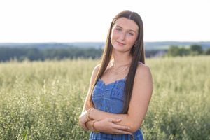 Young woman in blue dress stands in green field, long brown hair, arms crossed, soft golden hour light.