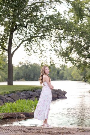Young woman in floral maxi dress smiling by a serene park lake surrounded by lush green trees.