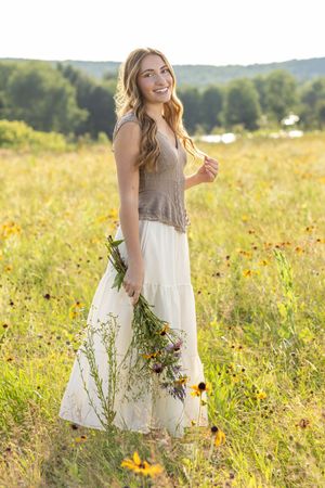 Young woman in white skirt holding wildflowers smiling in a sunny field of yellow flowers with green trees in background.