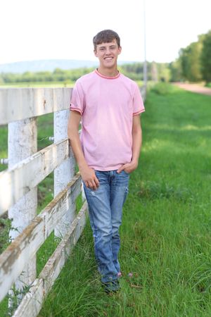Smiling teenage boy in pink t-shirt and jeans leaning against a white wooden fence in a green rural field.