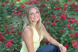 Young blonde woman smiling in yellow top and jeans, seated outdoors among vibrant red wildflowers.