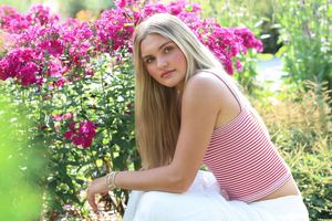 Young blonde woman in striped crop top and white skirt posing near vibrant pink bougainvillea flowers outdoors.