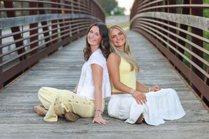 Two smiling young women sitting back-to-back on a wooden bridge, wearing white and yellow outfits on a sunny day.