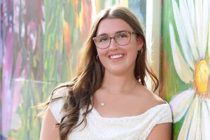 Smiling young woman with glasses and long brown hair poses in front of a colorful floral mural.