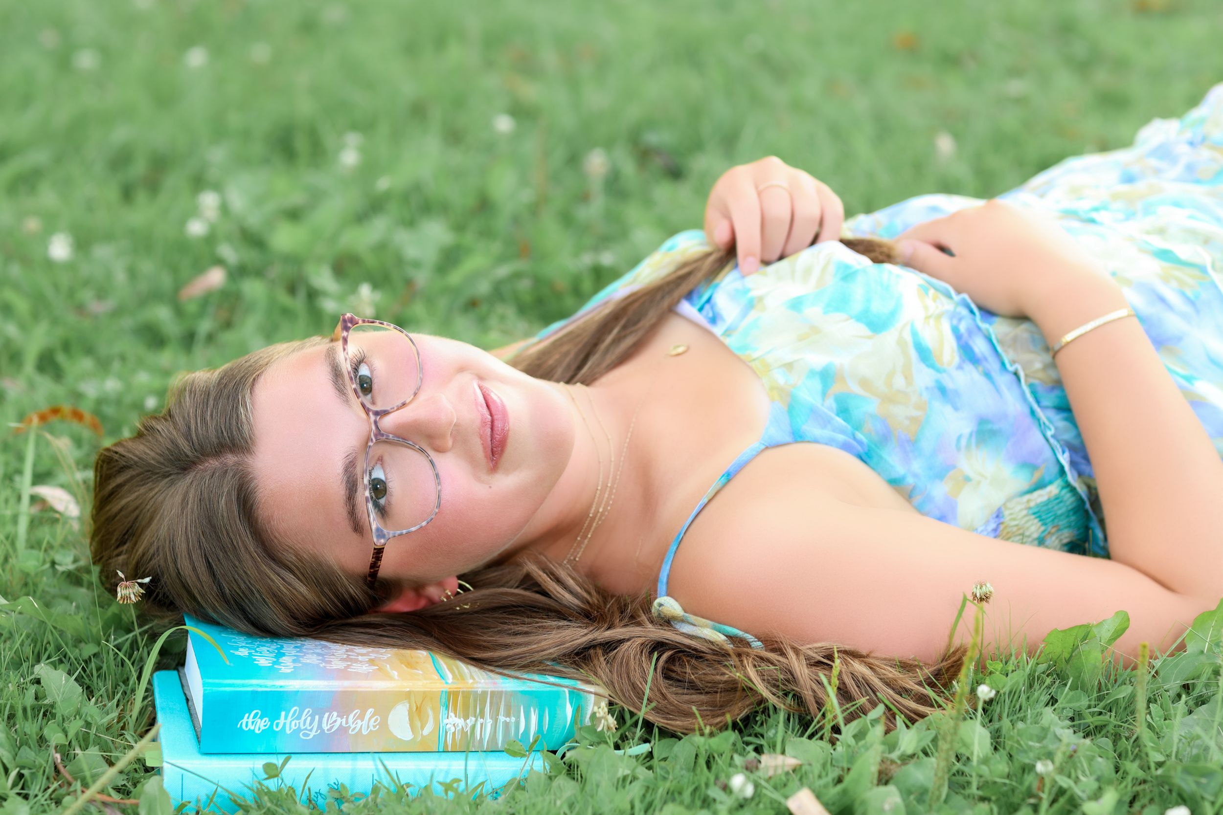 Young woman with glasses lying on green grass, smiling while resting her head on a colorful book outdoors.