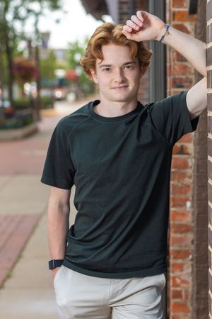 Young man with red hair in dark t-shirt leans against brick wall on urban sidewalk, smiling confidently.