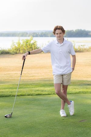 Young man in white polo and khaki shorts leaning on a golf club on a lush green golf course near a lake.