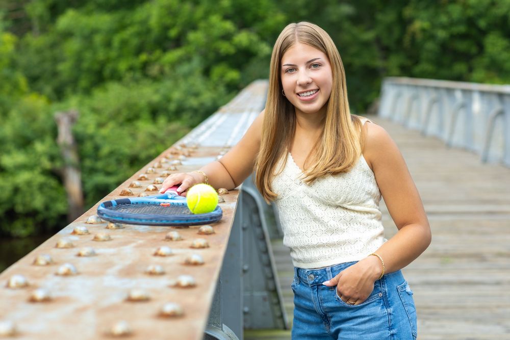 Smiling teenage girl holding a tennis ball on a rustic bridge, wearing a white top and denim shorts outdoors.