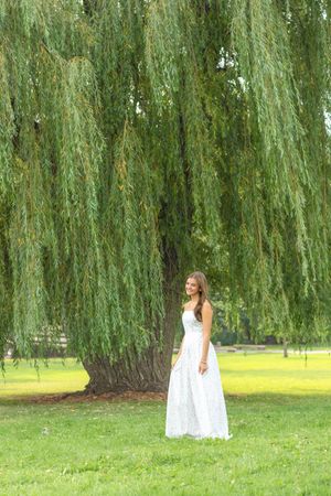 Young woman in white maxi dress standing under a weeping willow tree on lush green grass.