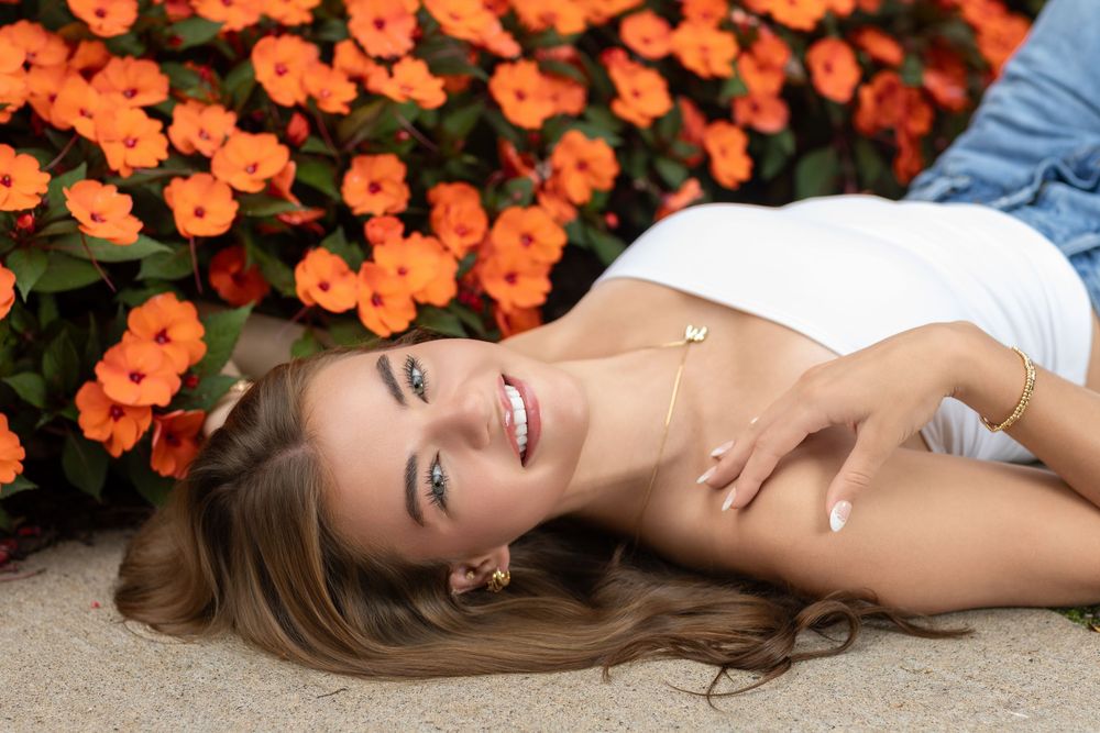 Young woman smiling while lying beside vibrant orange flowers in a white top and denim jacket outdoors.