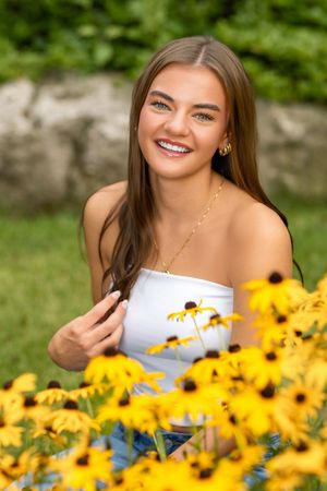 Smiling teenage girl with braces wearing white top posing among yellow black-eyed Susan flowers in a garden.