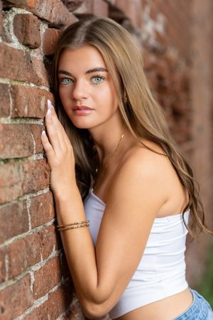 Young woman with green eyes and long blonde hair leaning against a brick wall wearing a white tube top and gold jewelry.