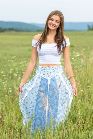Young woman in white crop top and blue floral maxi skirt smiling in a green meadow field.