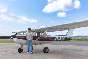 A person stands proudly beside a small white Cessna plane with tail number N11454 on a sunny airfield.