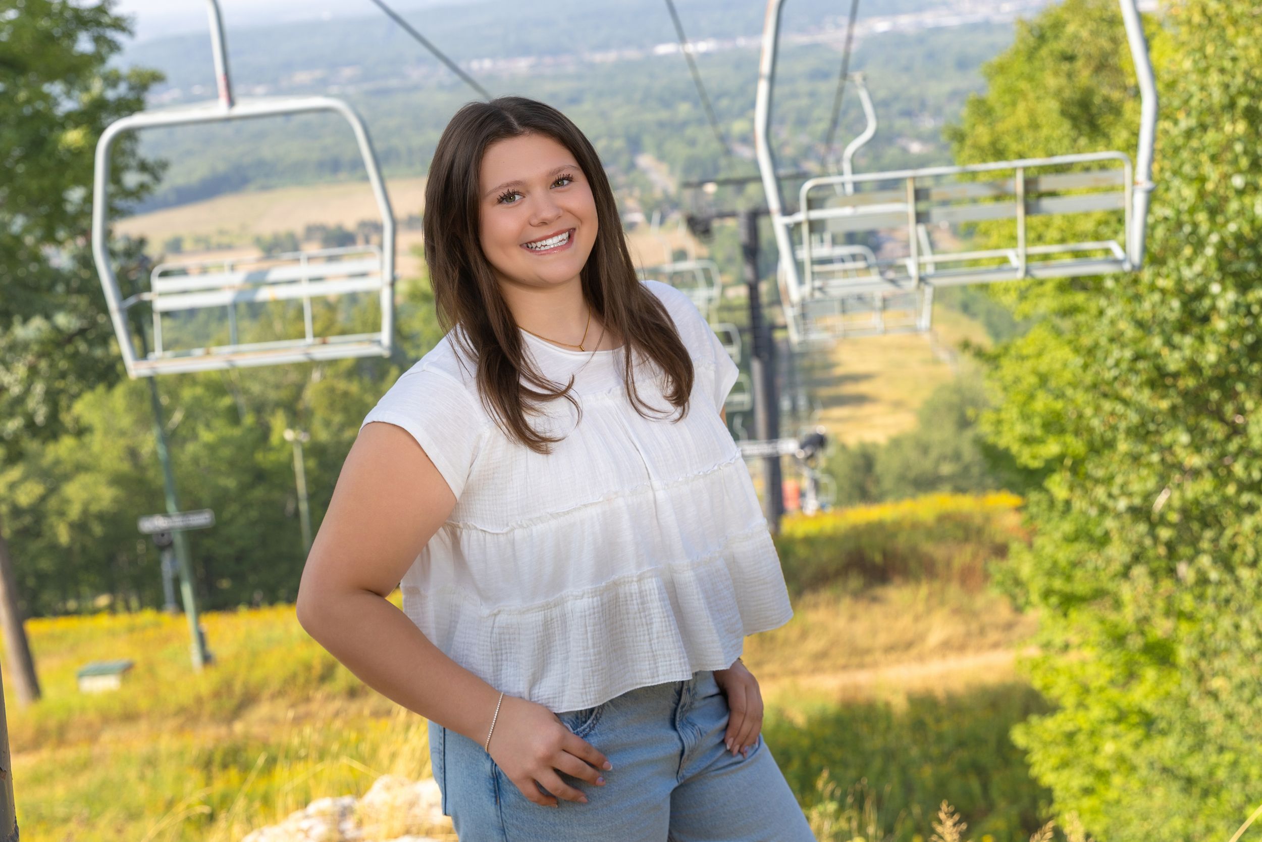 Young woman smiling in white top and jeans near ski chairlift with scenic green hills in background.