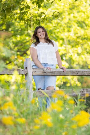 Smiling young woman in white top and jeans leaning on rustic wooden fence surrounded by lush green trees and yellow wildflowers.