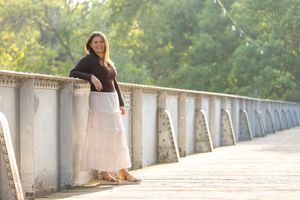 A young woman in a brown top and pink maxi skirt leans on a bridge railing surrounded by lush green trees.