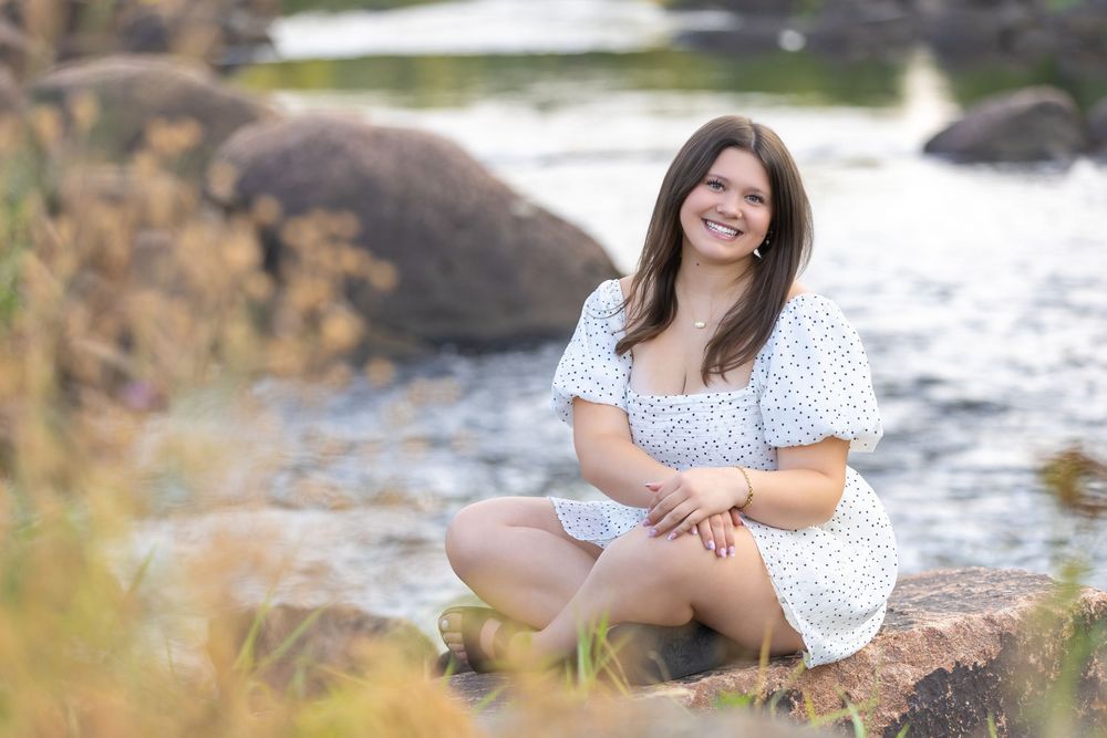 Young woman in floral dress smiling outdoors by rocky stream, surrounded by golden grasses in natural setting.
