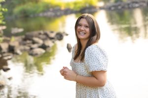 Young woman in white floral dress smiling outdoors near a reflective pond with rocks and greenery.