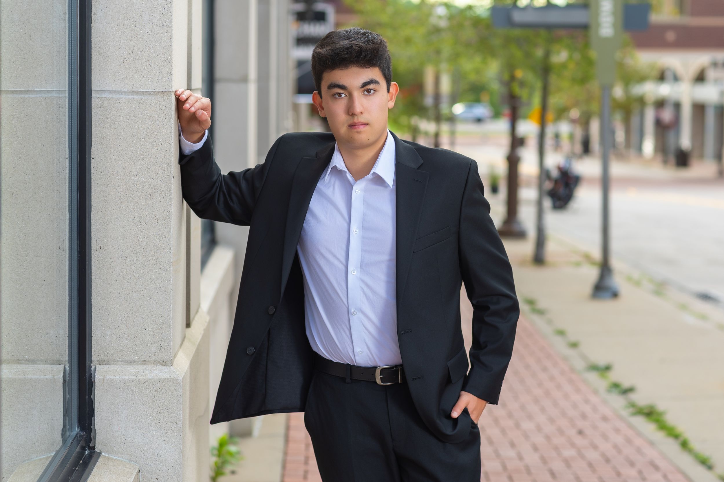 Young man in black suit posing confidently on urban sidewalk, leaning against building wall.