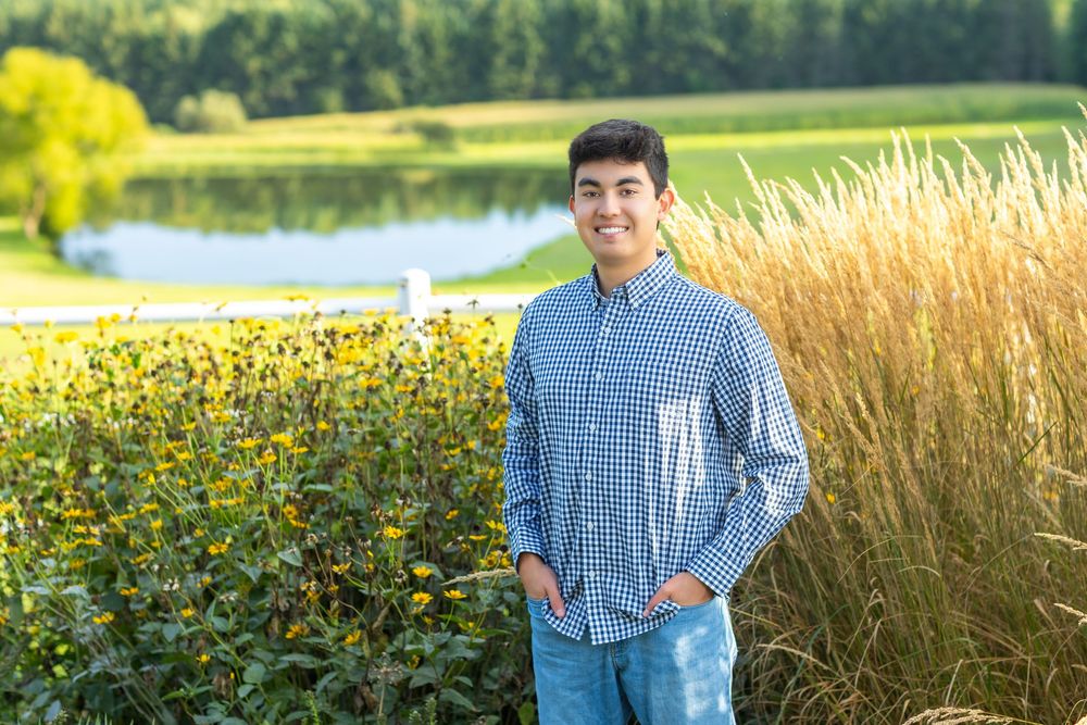 Young man in plaid shirt and jeans smiling outdoors near wildflowers and a pond on a sunny day.