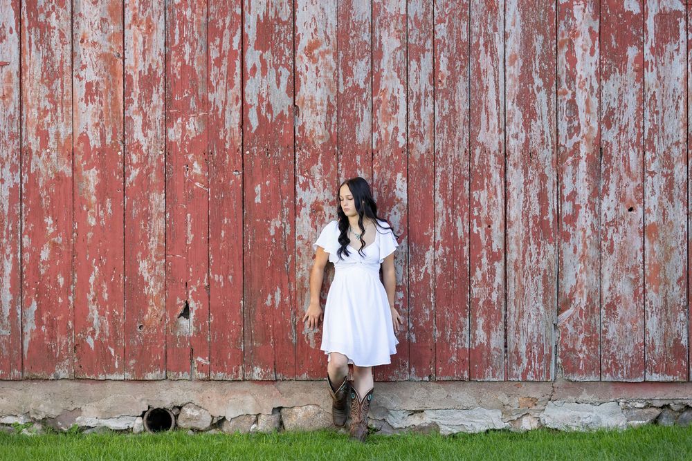 Young woman in white dress and cowboy boots poses against a rustic red peeling barn wall on green grass.