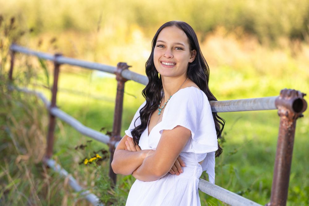 Young woman in white dress smiling with arms crossed, leaning against rustic fence in green field.