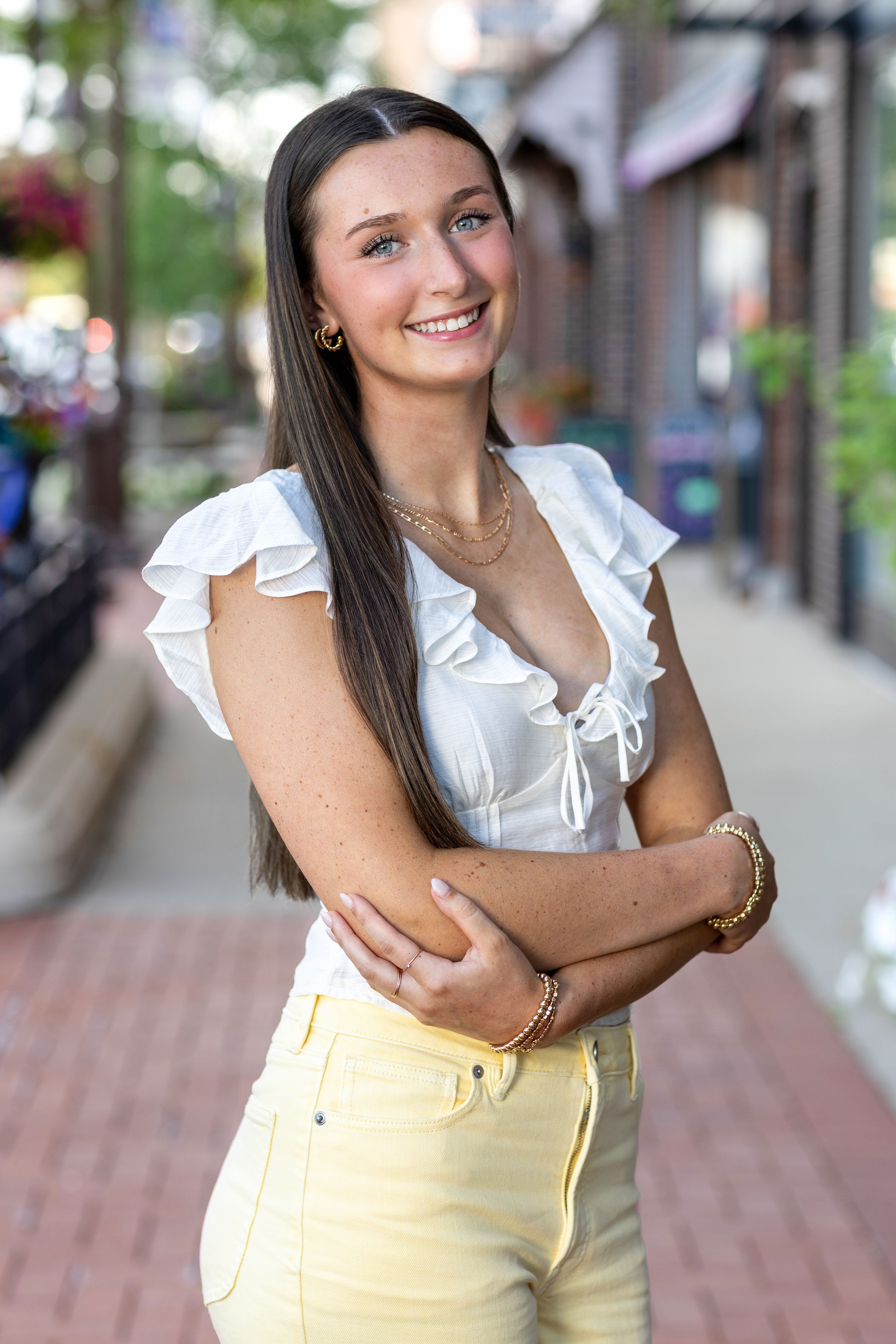 Young woman in white ruffled top and yellow jeans smiling outdoors on a brick sidewalk.