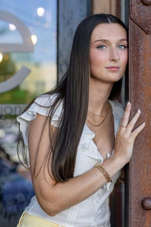 Young woman with long dark hair in white ruffled top leans against rustic metal door frame outdoors.