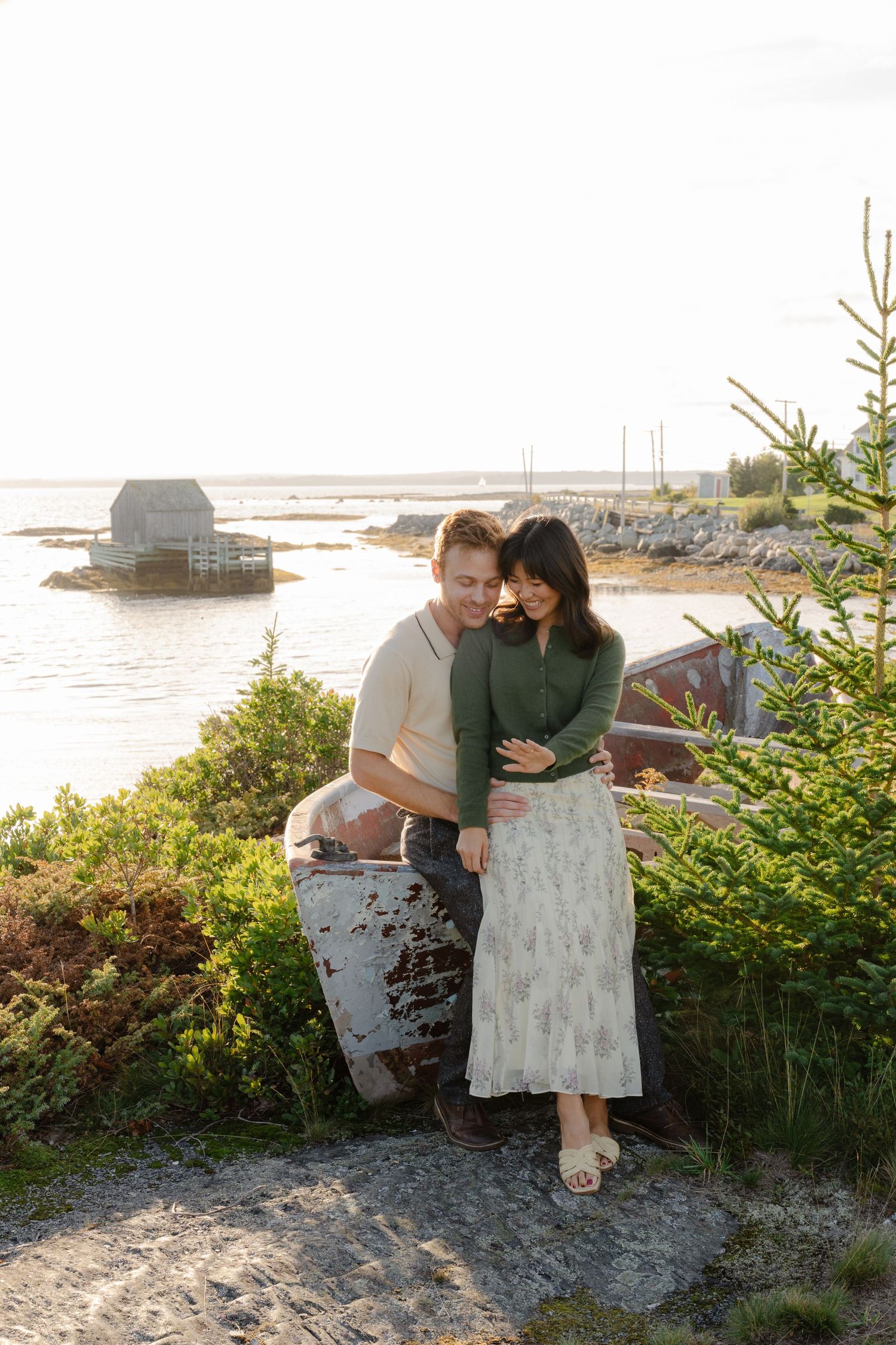 Couple embracing near coastal shoreline with rustic boat and fishing shack at golden hour.