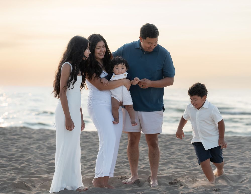 Family of five enjoying a sunset beach photoshoot, parents and three children posing on sandy shore in casual attire.