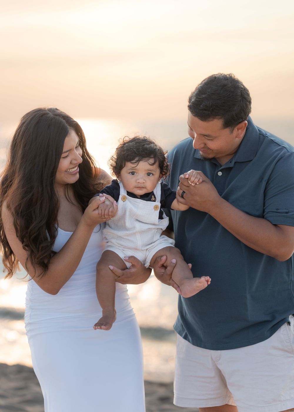 Family of three enjoying a golden sunset beach moment, parents lovingly holding their baby boy in white outfit.