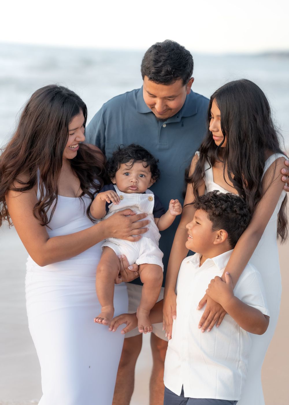 Family of five smiling together on a beach, parents holding a baby while two children gather around them.