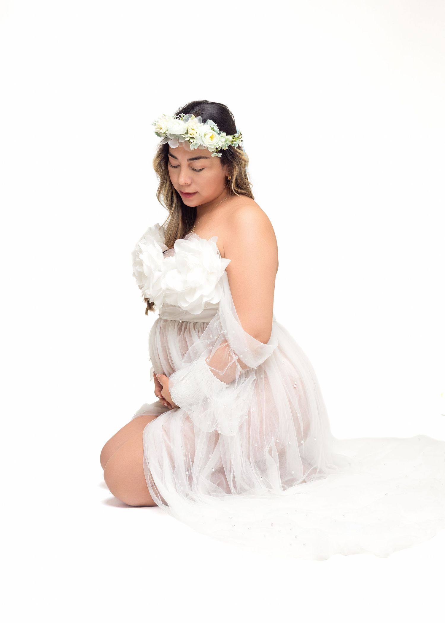 Pregnant woman in white tulle dress and floral crown kneels gracefully, cradling white flowers in a maternity photo.