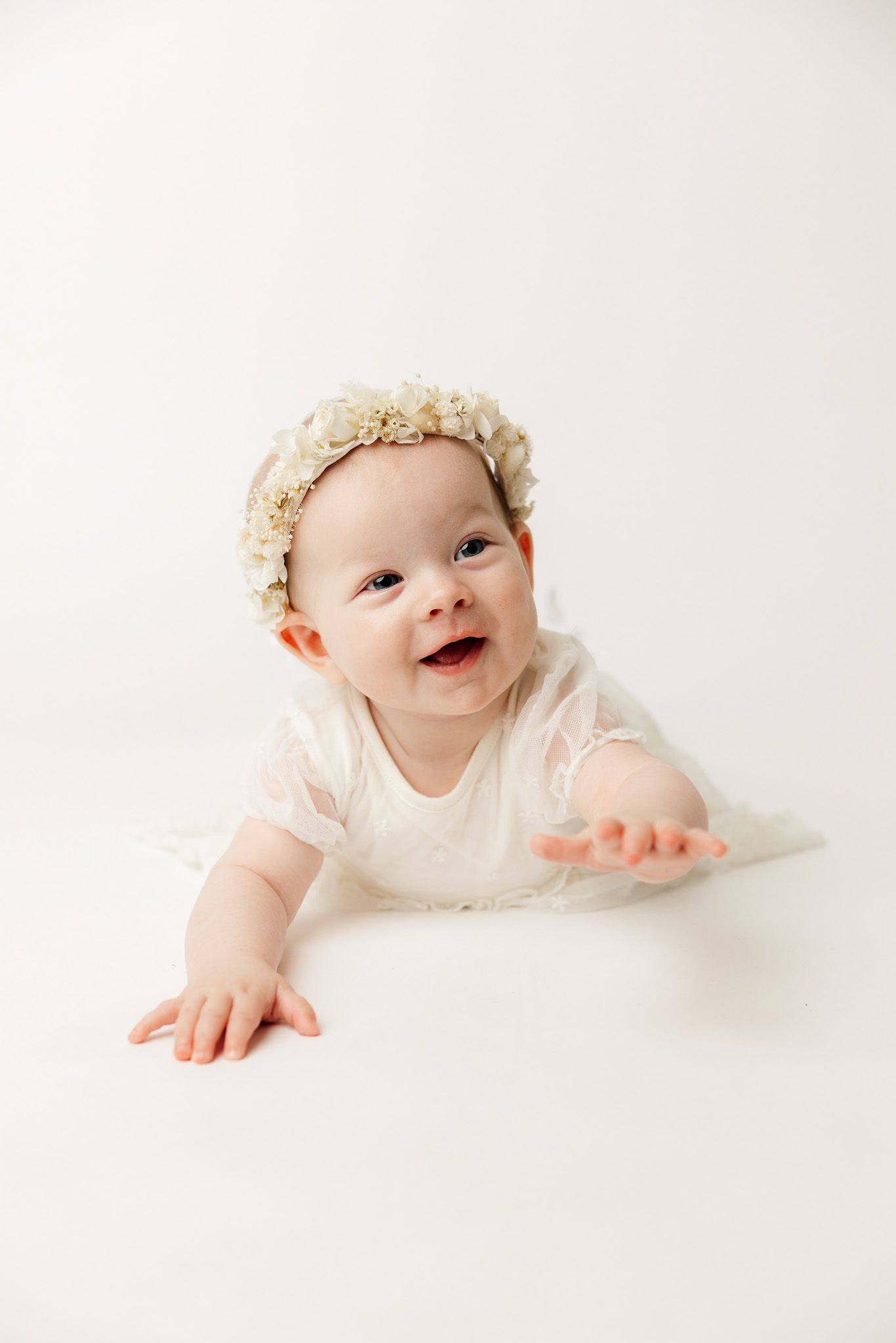 Adorable baby girl in white lace romper and floral crown sitting on white background.