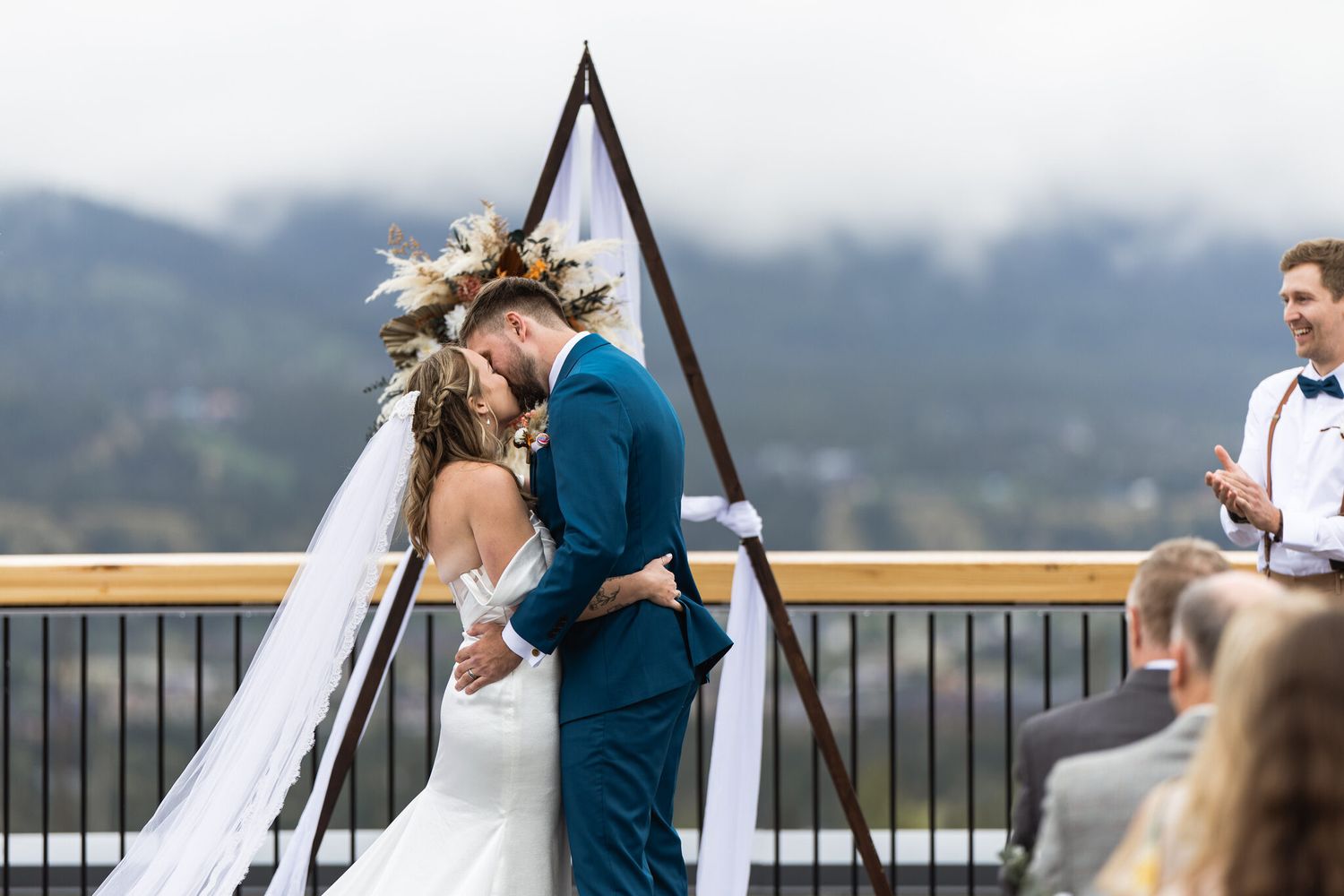 Bride and groom first kiss under floral arbor with clouded mountains at Vista Canmore