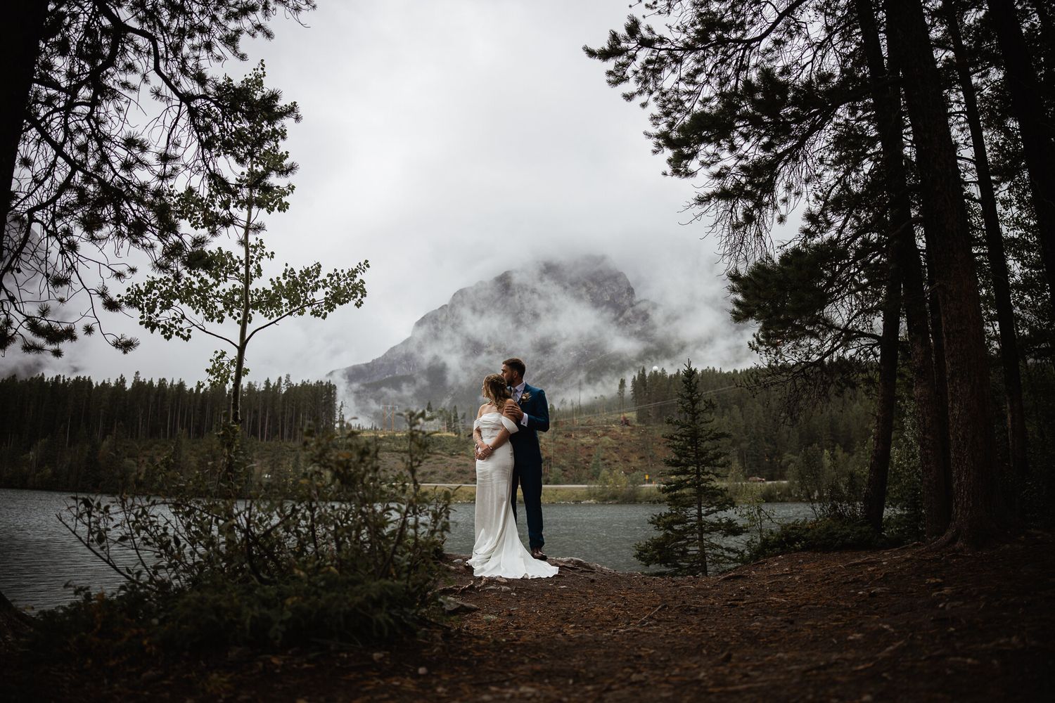 Bride and groom by a lake framed by pines with a misty mountain behind, Canmore wedding
