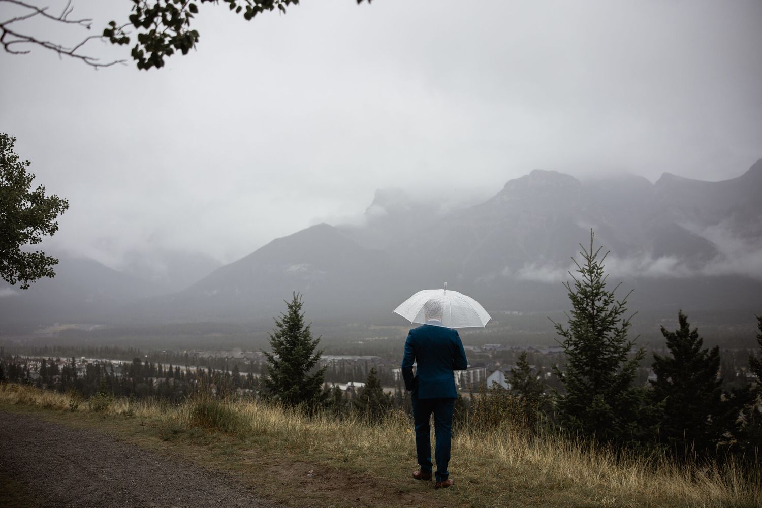 Groom in navy suit with clear umbrella waiting for first look at Vista Canmore, misty Rocky Mountains