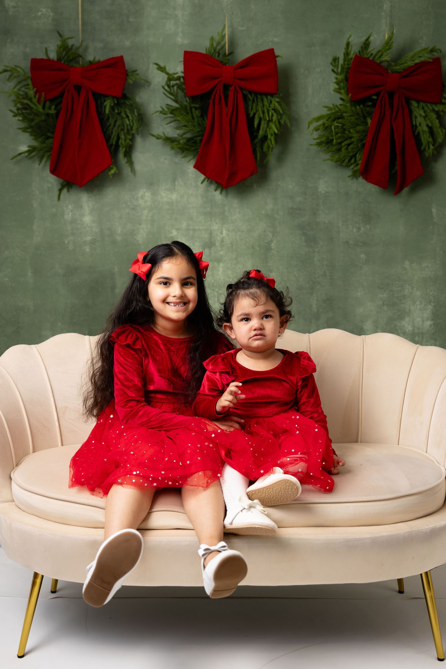 Two young girls in matching red dresses sit on a cream sofa with Christmas greenery and red bows on a green wall.