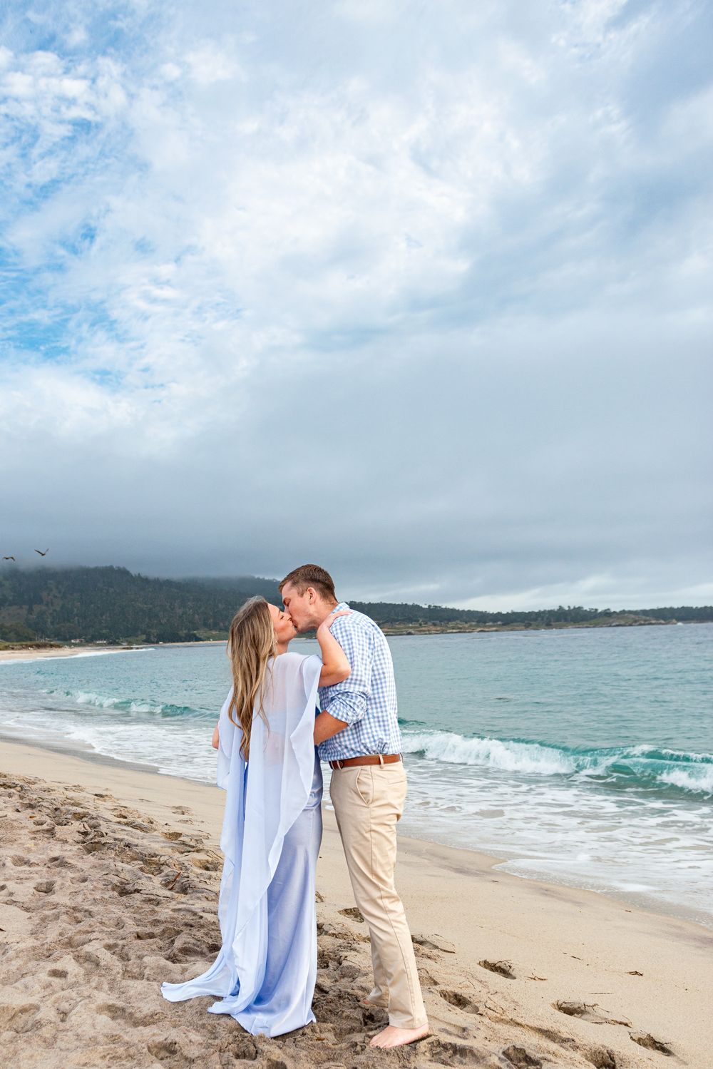Couple in light blue and khaki outfits kiss on a sandy beach with gentle waves and a cloudy blue sky behind them.