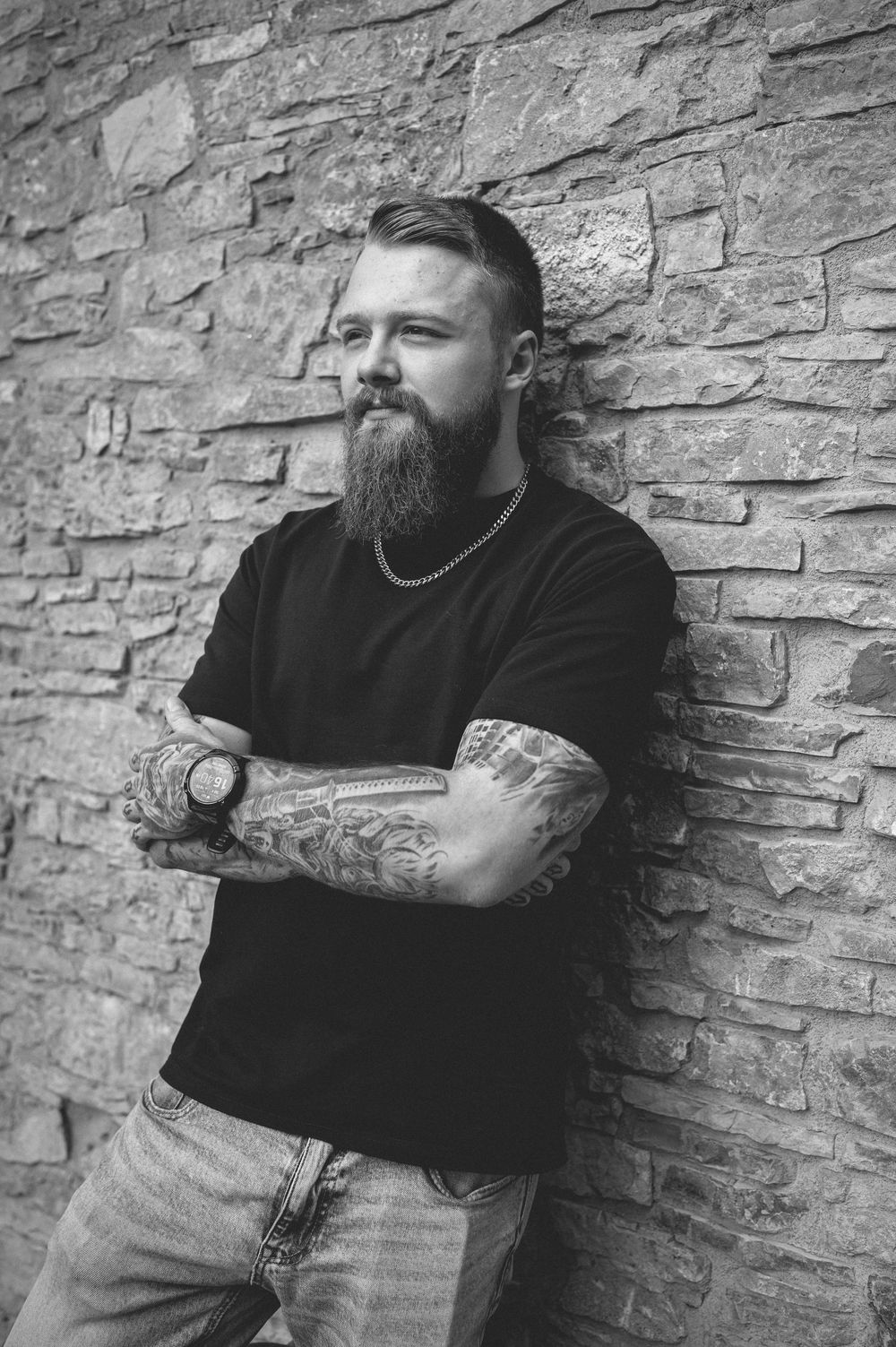 Bearded man with tattooed forearms and a chain necklace leans against a rustic stone wall in a black and white portrait.
