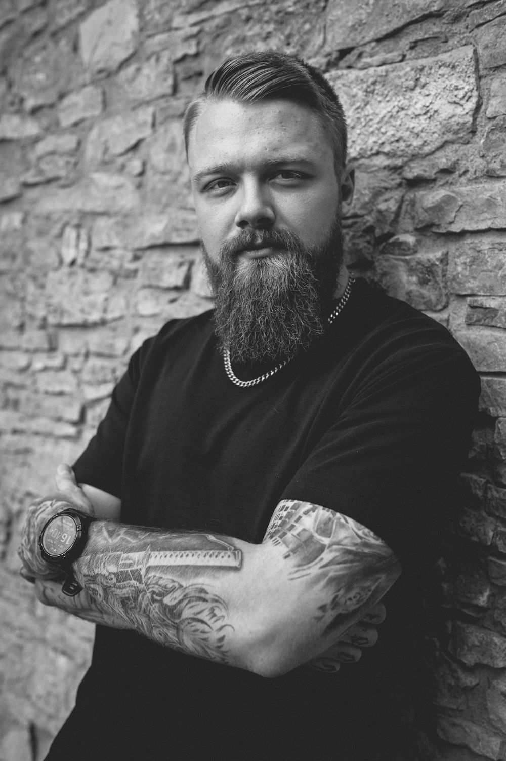 Black and white portrait of a bearded man with tattooed forearms leaning against a stone wall in a black t-shirt and chain necklace.