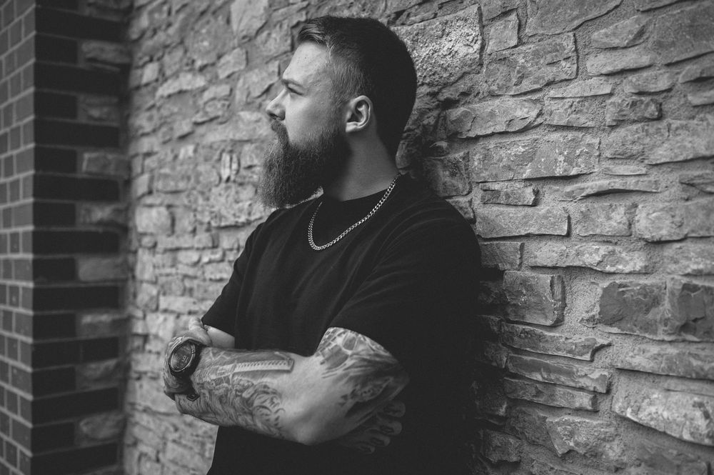 Bearded man with tattooed forearms and a chain necklace leans against a rustic stone brick wall, gazing to the side in black and white.
