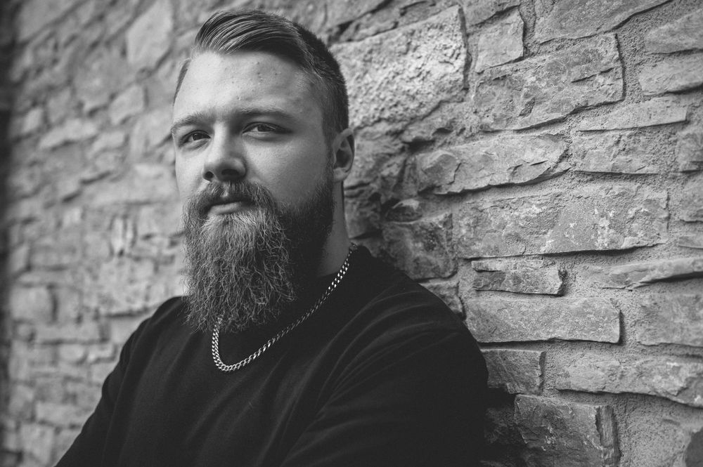 Black and white close portrait of a bearded man in a dark sweater and chain necklace leaning against a rustic stone brick wall.