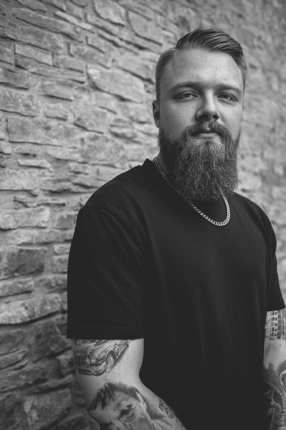Black and white portrait of a bearded man with tattooed arms and a chain necklace standing against a stone wall.