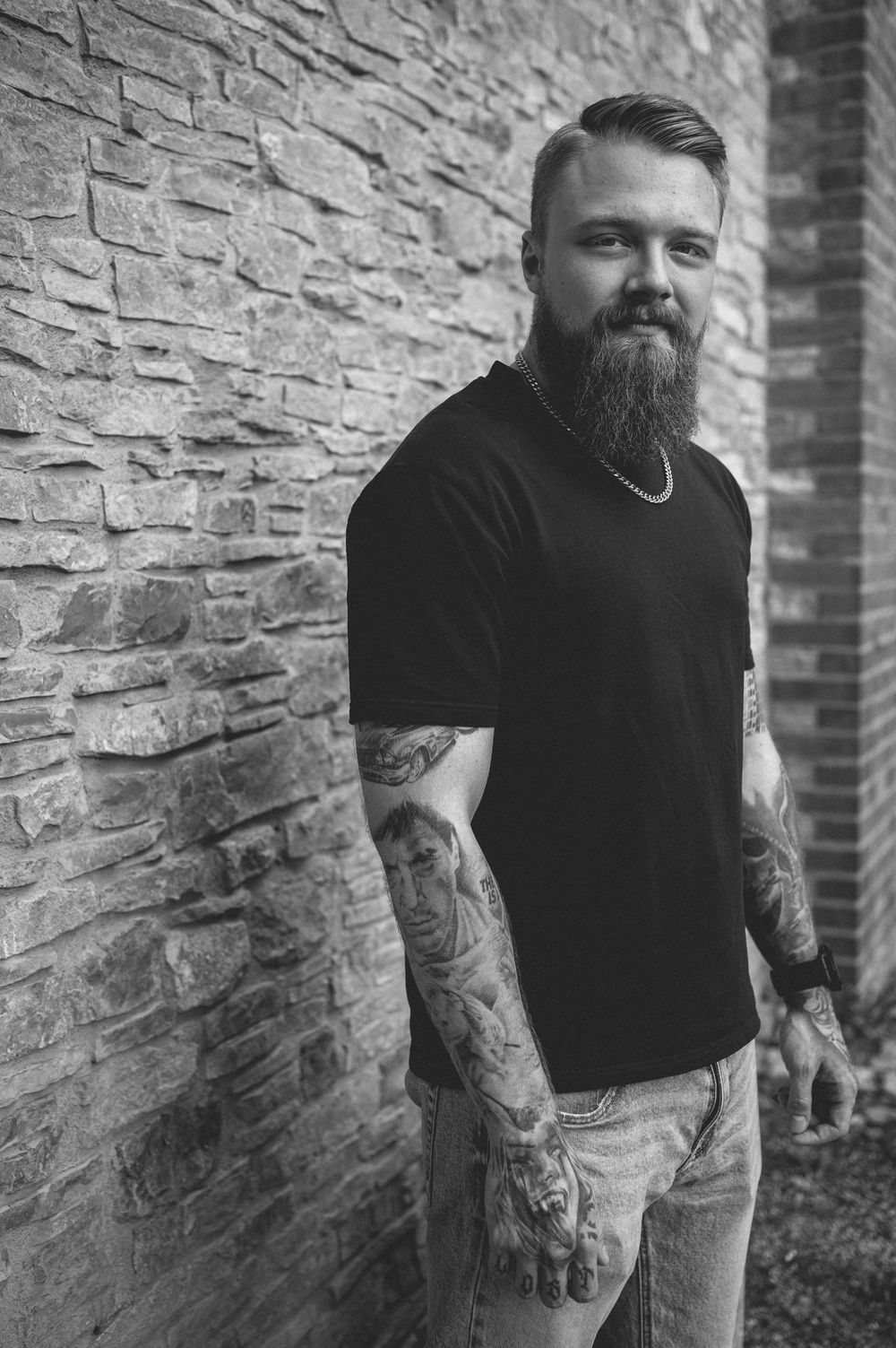Black and white photo of a bearded man with arm tattoos, wearing a black t-shirt and chain necklace, standing against a stone wall.