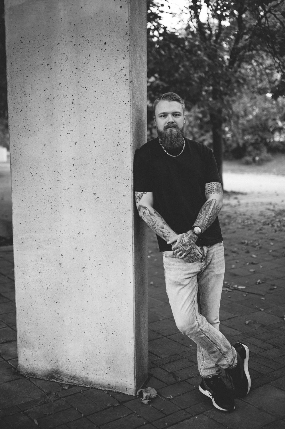 Black and white portrait of a bearded, tattooed man in a dark t-shirt and light jeans leaning against a concrete pillar outdoors.